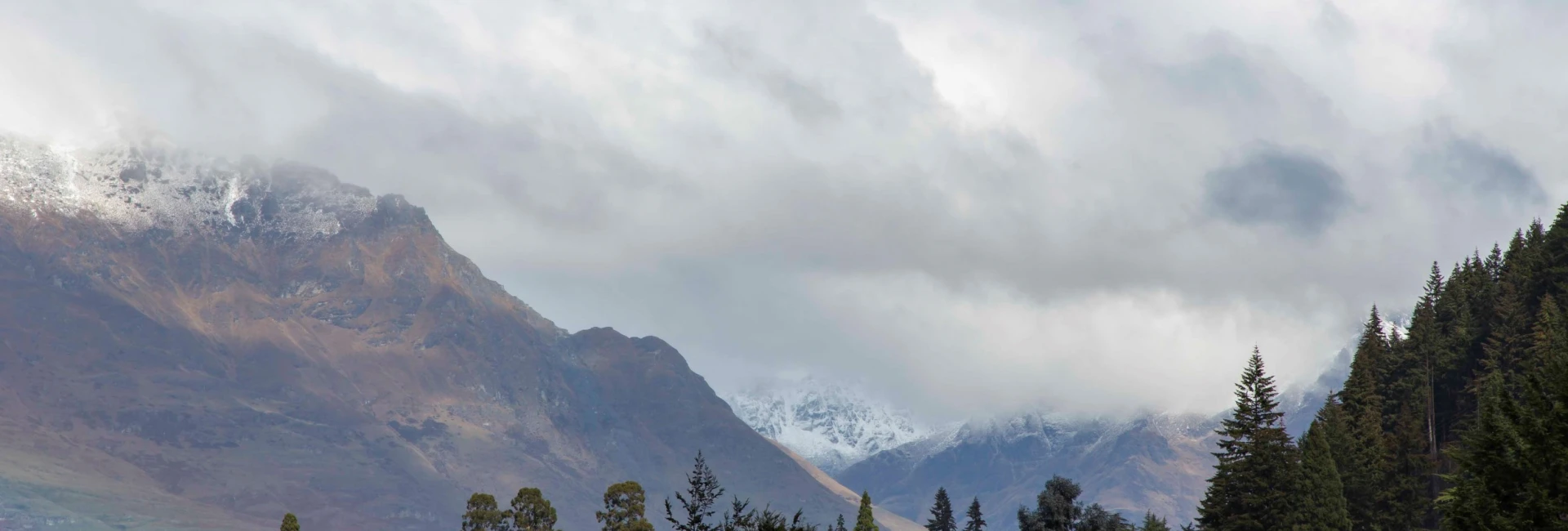 A moody mountain scene with clouds, trees, and snow.